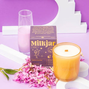 White candle in a yellow glass container in front of a purple packaging box with 'Milkjar' branding. Both items sit on a purple and white surface with flowers. 