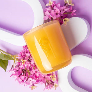 Yellow candle container on a white and purple surface with flowers in the background.