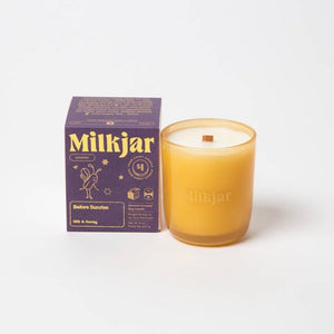 White candle in a yellow glass container shown next to its purple packaging box on a white background.