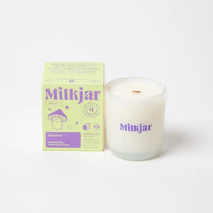 Canadian company Milkjar candle and its box packaging. The white candle is in a translucid glass with "Milkjar" written in purple. The other side of the candle has a cute smiling mushroom doodle in purple. The packaging is a small lime-coloured box with purple text and graphics. 