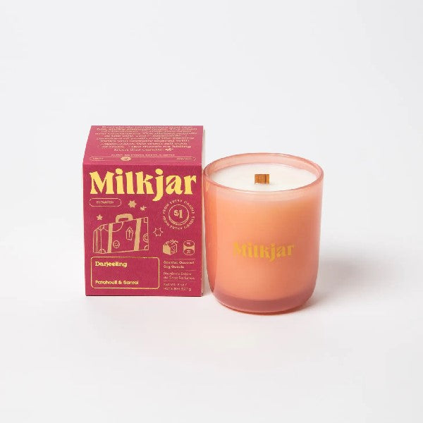 Canadian company Milkjar candle and its box packaging. The white candle is in an orange glass with "Milkjar" written in yellow. The other side of the candle has a cute briefcase doodle in yellow. The packaging is a small red box with yellow text and graphics. 