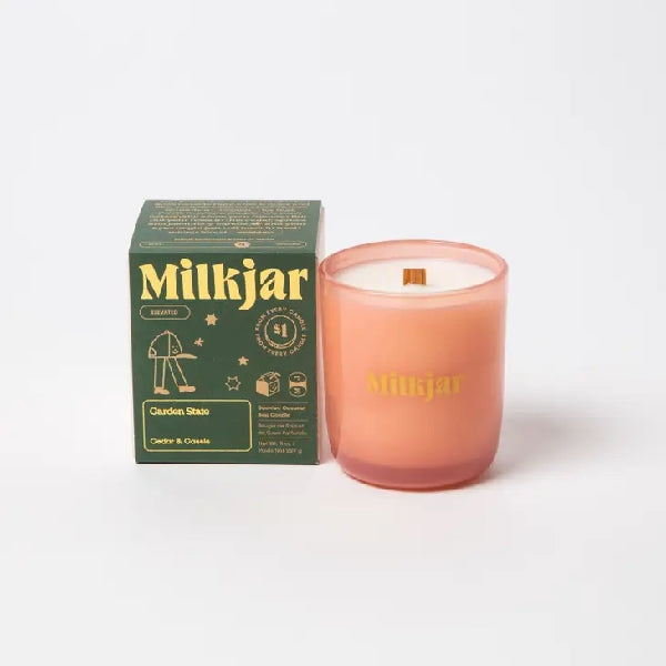 Canadian company Milkjar candle and its box packaging. The white candle is in an orange glass with "Milkjar" written in yellow. The other side of the candle has a cute smiling baseball hat with legs doodle in yellow. The packaging is a small green box with yellow text and graphics. 