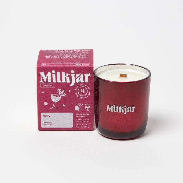 Canadian company Milkjar candle and its box packaging. The white candle is in a red glass with "Milkjar" written in grey. The other side of the candle has a cute smiling glass with holly doodle in grey. The packaging is a small red box with grey text and graphics. 