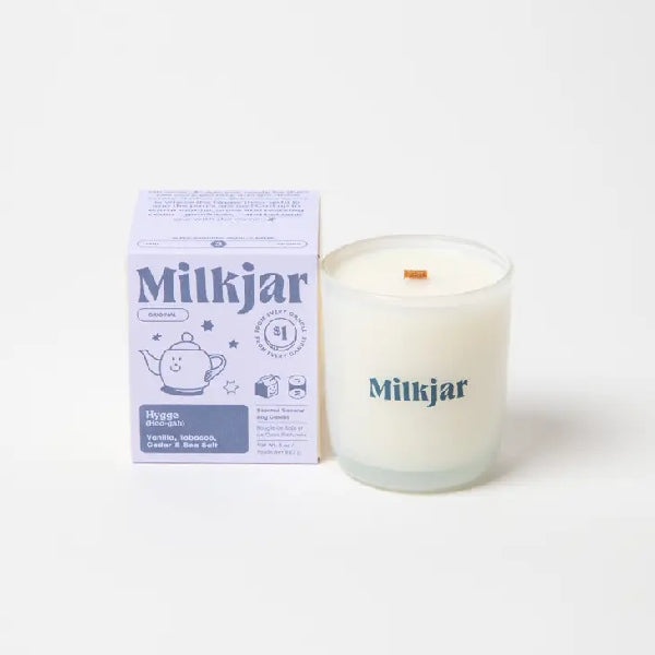 Canadian company Milkjar candle and its box packaging. The white candle is in a translucid glass with "Milkjar" written in dusty purple. The other side of the candle has a cute smiling teapot doodle in dusty purple. The packaging is a small lilac box with dusty purple text and graphics. 