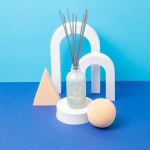 Decorative diffuser containing reed sticks on a white stand placed in front of white arch-shapes against a blue background