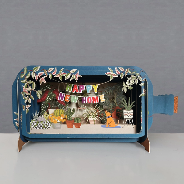 blue bottle pop up card. the intricate paper cuttings depict a homey scene with plants, a dog, and an rainbow hanging banner that reads "happy new home"